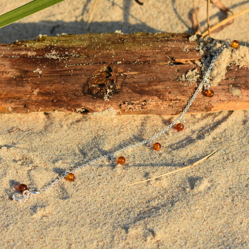 Srebrna bransoletka z brązowymi bursztynami leżąca na plaży na kawałku drewna.