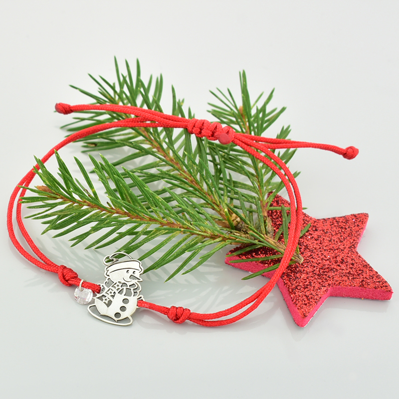 Bransoletka świąteczna bałwanek srebrny na czerwonym sznurku leżąca na gałązce świerku.