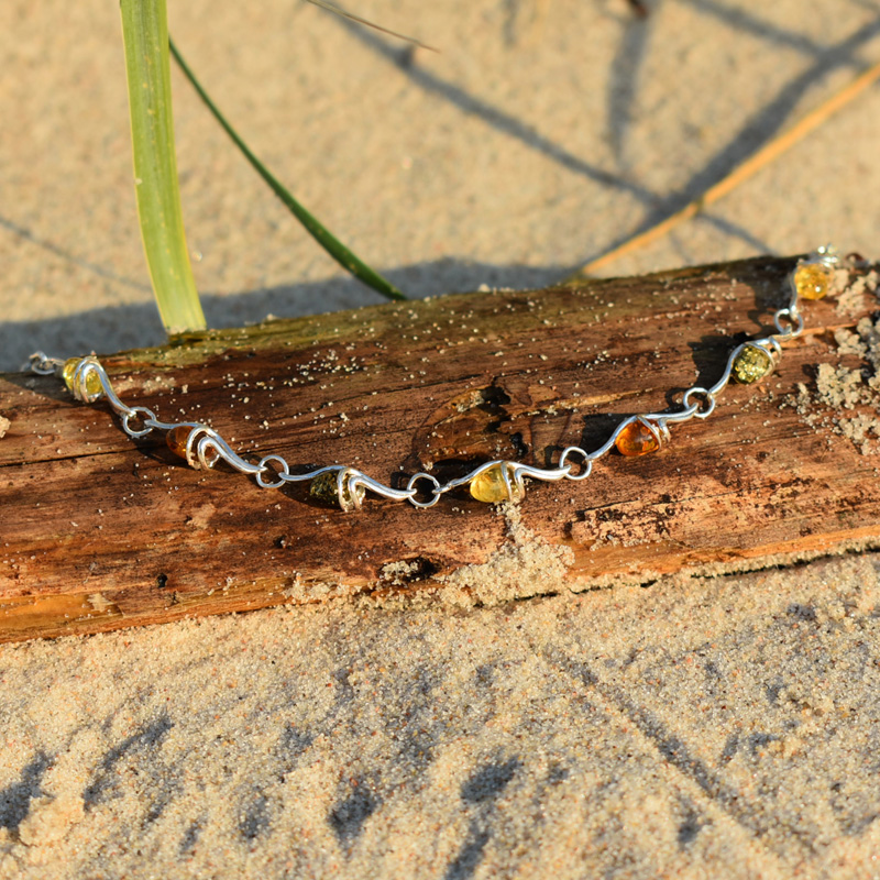 Srebrna bransoletka z 3 kolorami bursztynów - brązowym, zielonym i żółtym, leżąca na plaży na kawałku drewna.