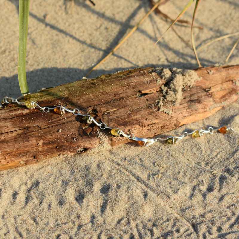 Srebrna bransoletka z 3 kolorami bursztynów - brązowym, żółtym i zielonym, leżąca na plaży na kawałku drewna.