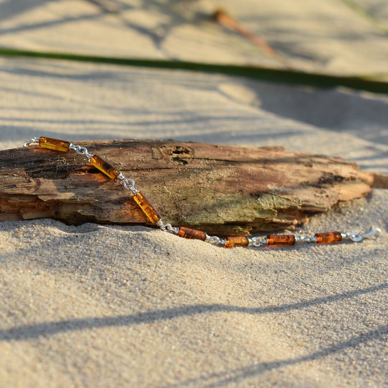Srebrna bransoletka z podłużnymi bursztynami w kolorze brązowym, leżąca na plaży na kawałku drewna.