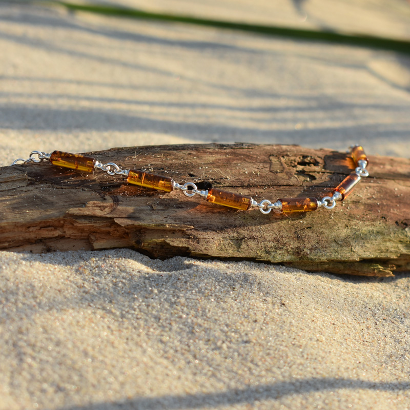 Srebrna bransoletka z podłużnymi bursztynami w kolorze brązowym, leżąca na plaży na kawałku drewna.