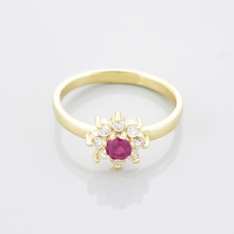 Złoty pierścionek kwiatek z białymi małymi cyrkoniami i jedną dużą, czerwono malinową po środku leżący przodem.
