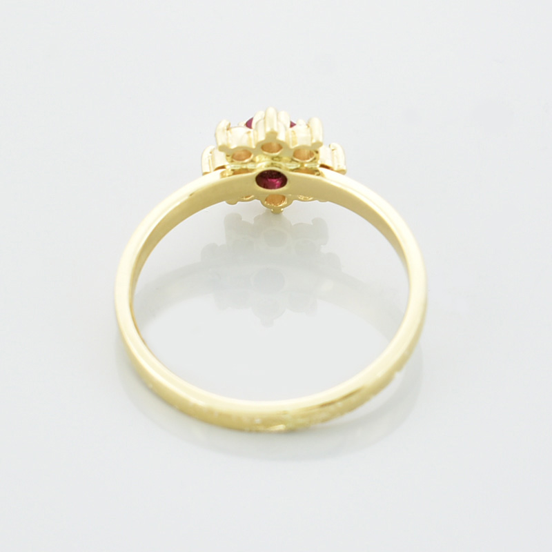 Złoty pierścionek kwiatek z białymi małymi cyrkoniami i jedną dużą, czerwono malinową po środku leżący tyłem.