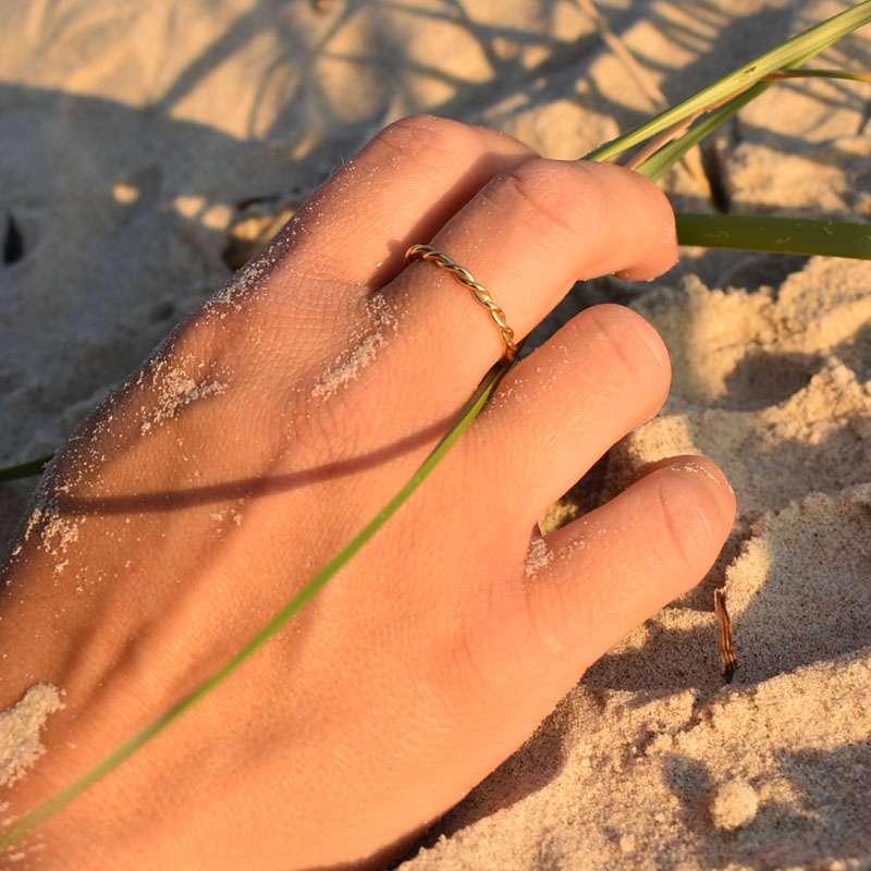 Pierścionek pleciony pozłacany przedstawiony na dłoni leżącej na piasku.