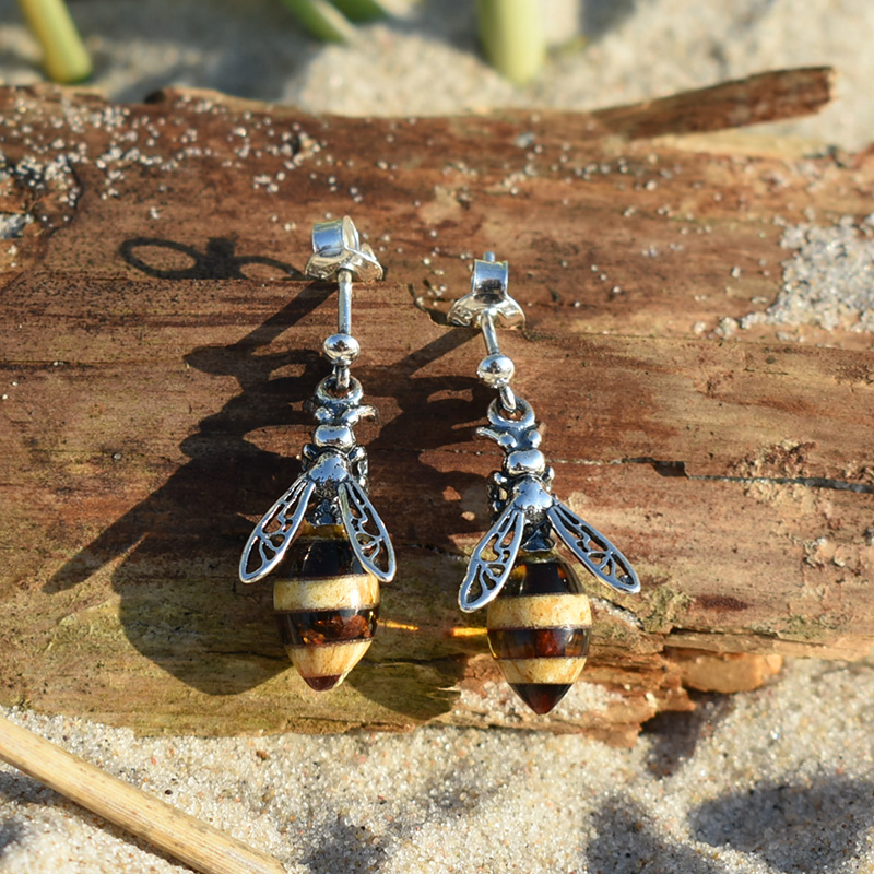 Kolczyki wiszące pszczoły z bursztynami bałtyckimi przedstawione na plaży na kawałku drewna.