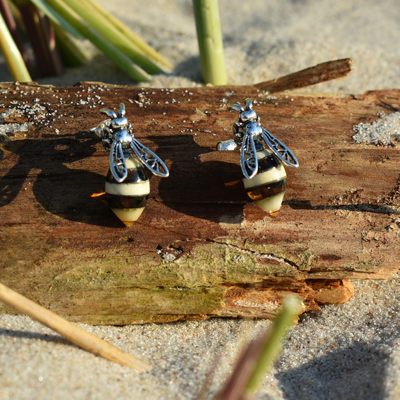 Kolczyki pszczoły na sztyfcie z bursztynami wykonane w srebrze, leżące na plaży na kawałku drewna.