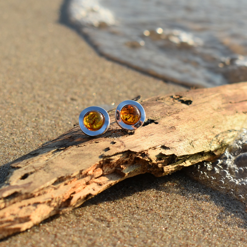Okrągłe srebrne kolczyki z bursztynem leżące na plaży na kawałku drewna.