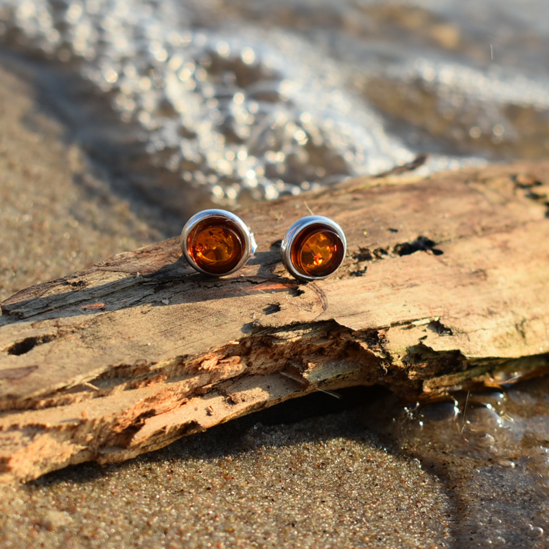Srebrne okrągłe kolczyki z brązowym bursztynem leżące na plaży na kawałku drewna.