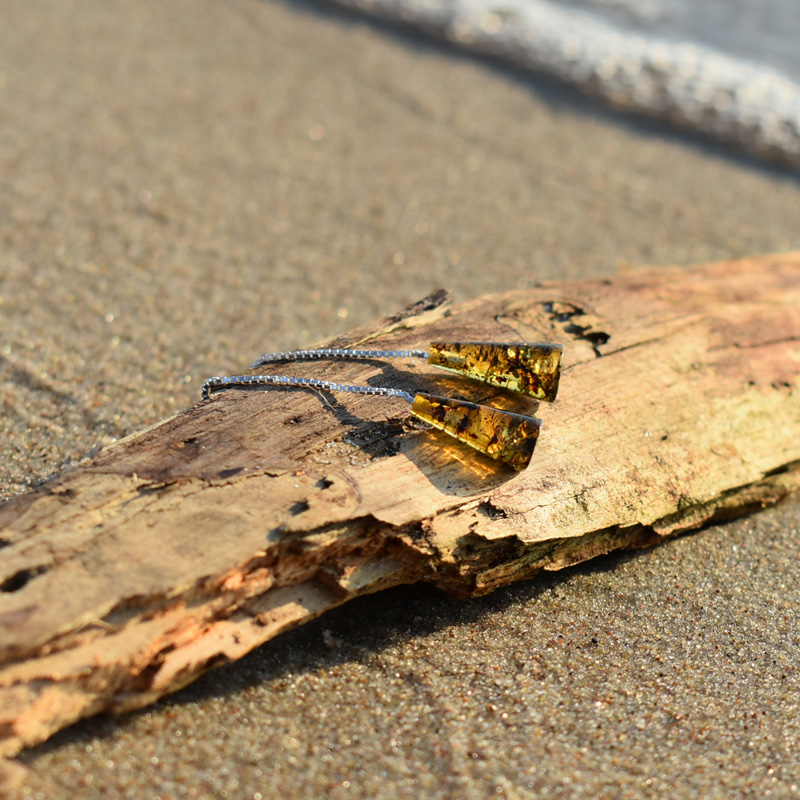 Kolczyki wiszące z zielonym bursztynem leżące na plaży na kawałku drewna.