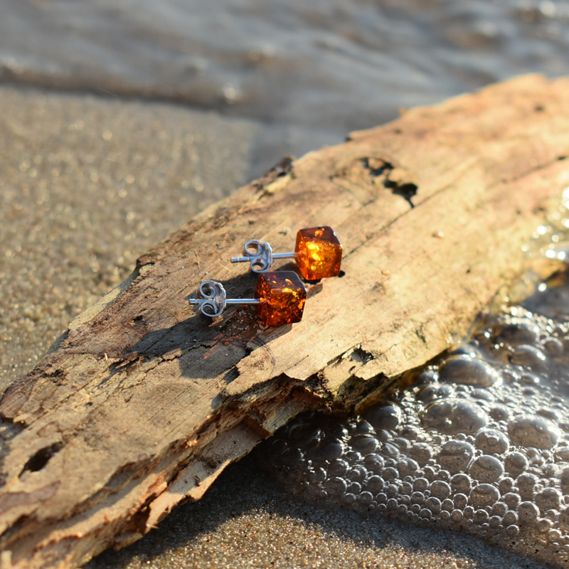 Kwadratowe kolczyki z bursztynem leżące na kawałku drewna na plaży.