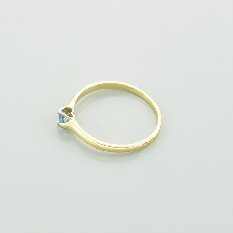 Złoty pierścionek serce z topazem w kolorze błękitnym leżący na lewym boku.