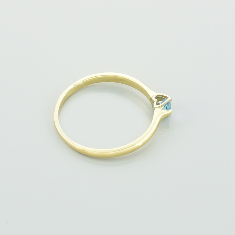 Złoty pierścionek serce z topazem w kolorze błękitnym leżący na prawym boku.