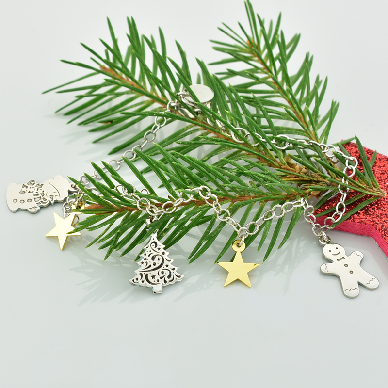 Srebrna bransoletka świąteczna z zawieszkami - bałwankiem, piernikowym ludzikiem, choinką i pozłacanymi gwiazdkami leżąca na gałązce świerku.