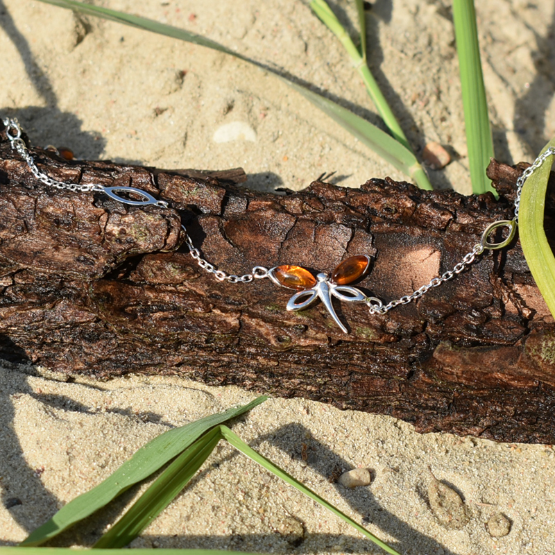 Srebrna bransoletka z bursztynami ważka na łańcuszku z regulacją długości przedstawiona na kawałku drewna na plaży.