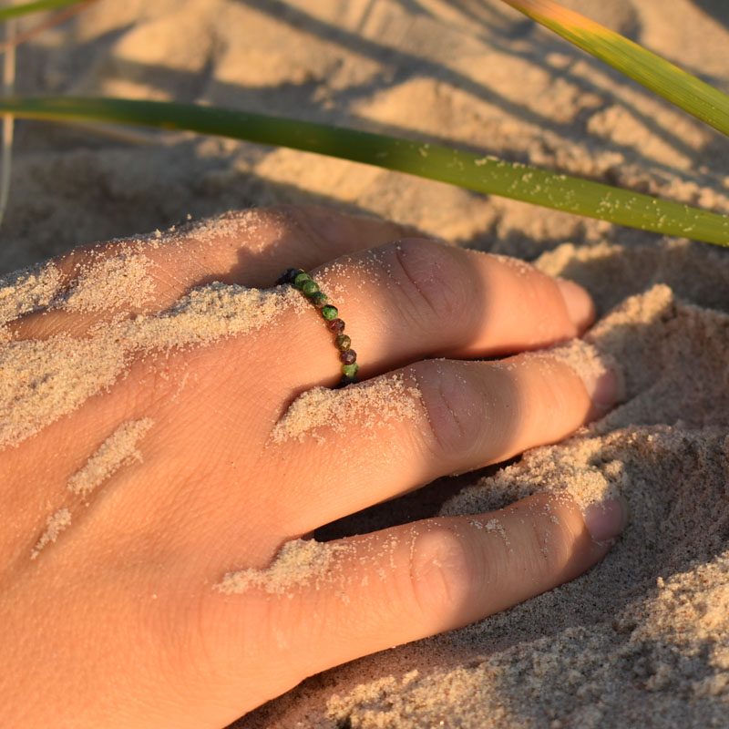 Pierścionek na gumce z okrągłych kamieni naturalnych - zoisytów i rubinów przedstawiony na dłoni leżącej w piasku.